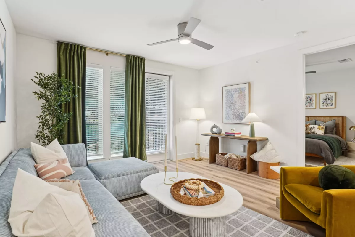Light-filled living room with green curtains, soft sectional, and open doorway to bedroom.