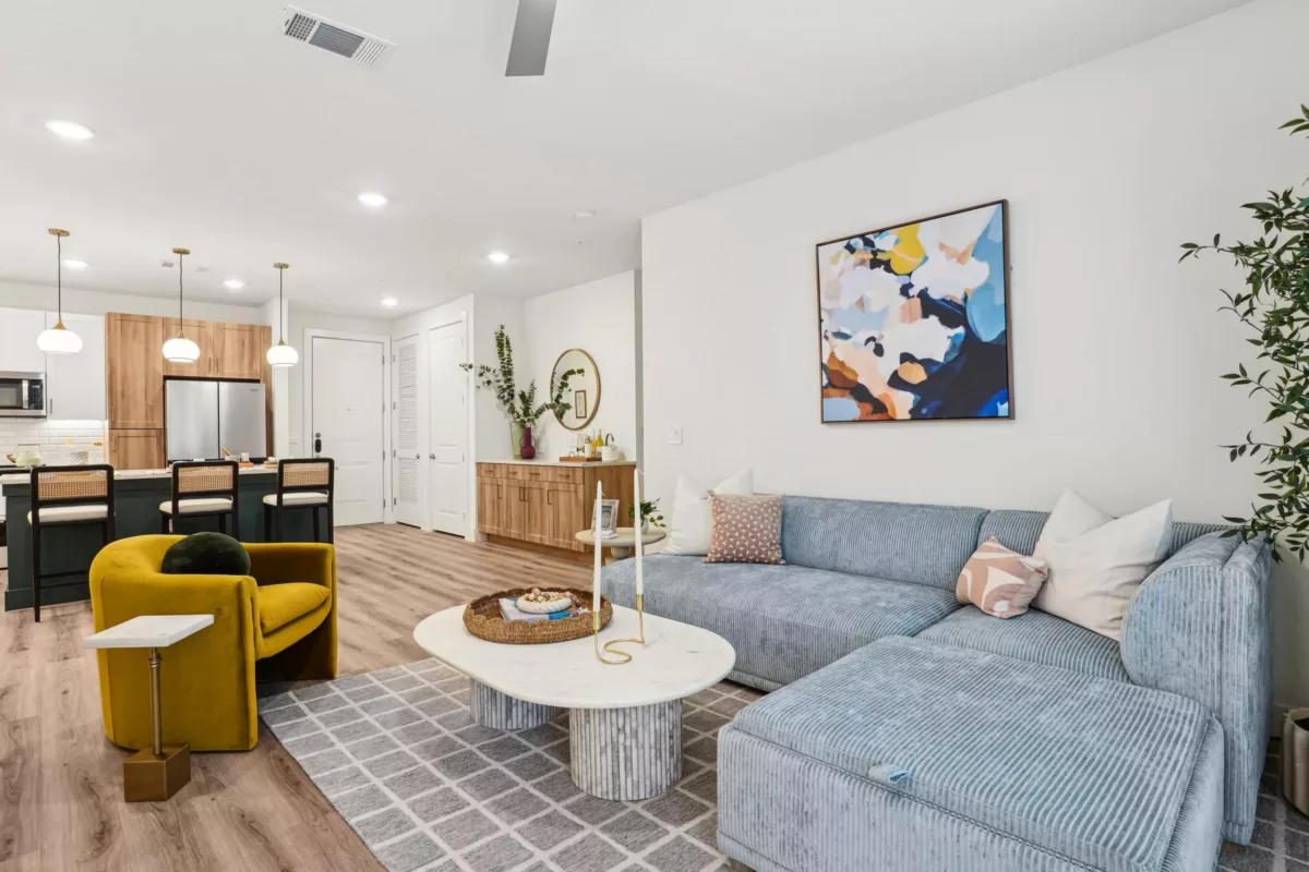 Cozy living room with blue sectional, yellow chair, and abstract art near open kitchen.